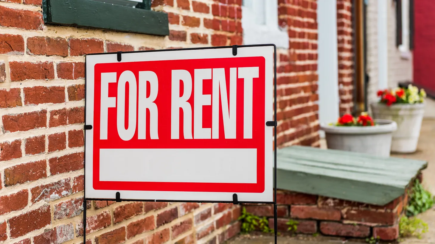 A “for rent” sign is erected in front of a brick building.
