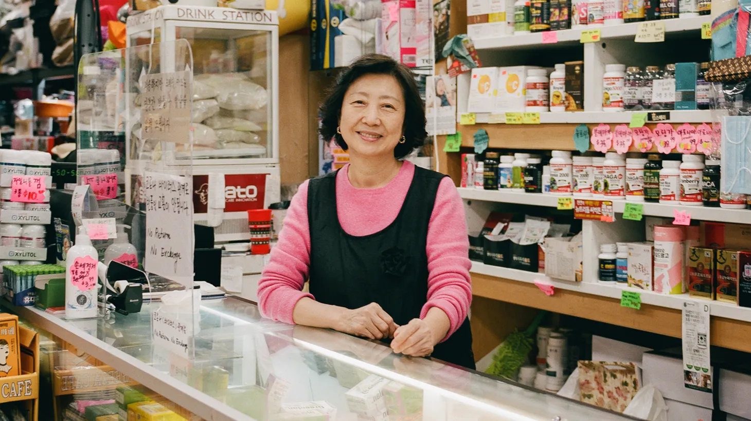 Ms. Jo, shopkeeper at Home Mart Plus Co. in Koreatown, stands behind a glass counter wearing a pink sweater and black vest.