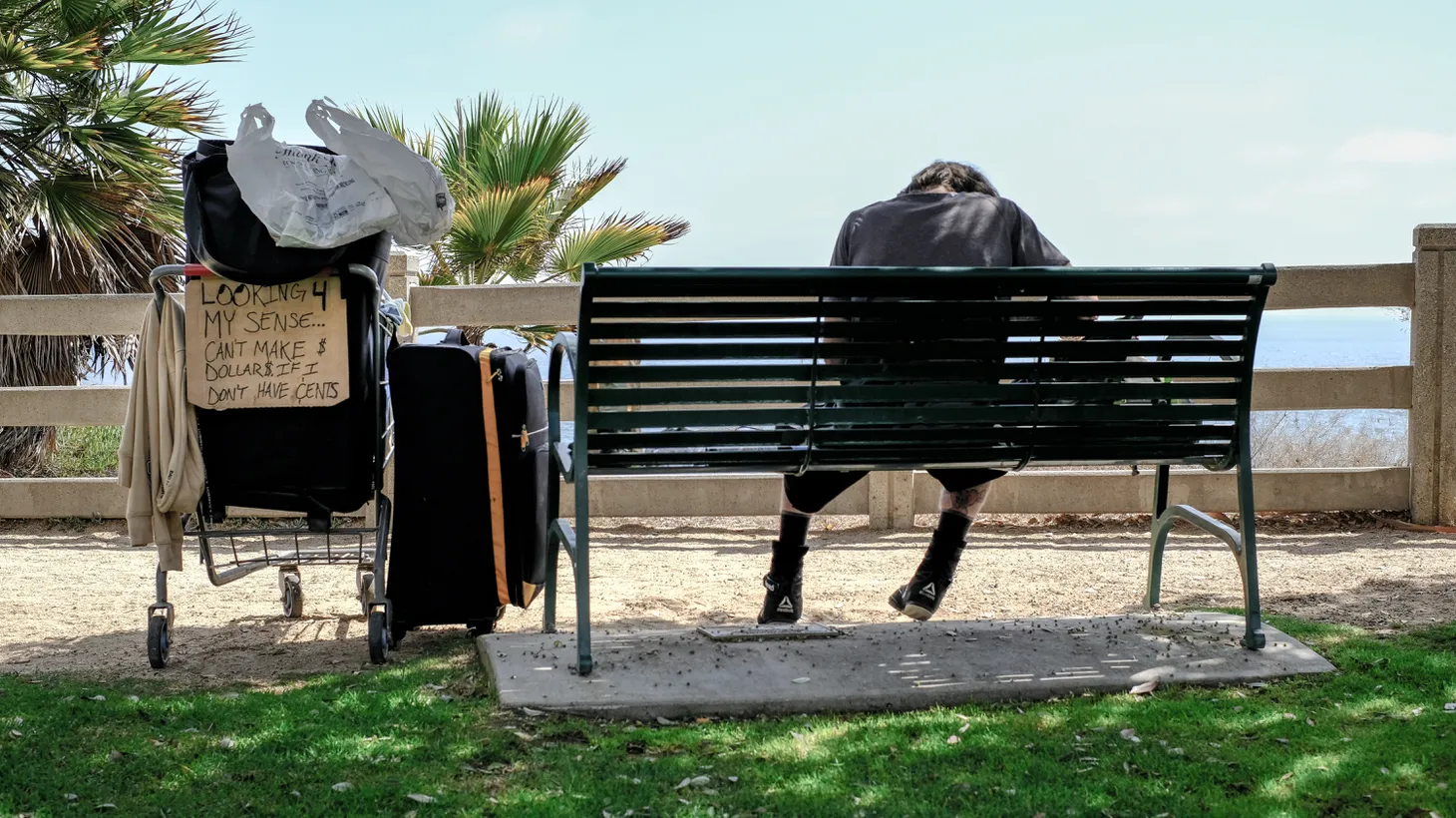 An unhoused person sits on a bench in Palisades Park in Santa Monica. His cart of possessions includes a sign that says, “Looking 4 my sense … can’t make $ dollars $ if I don’t have cents.” July 25, 2022.