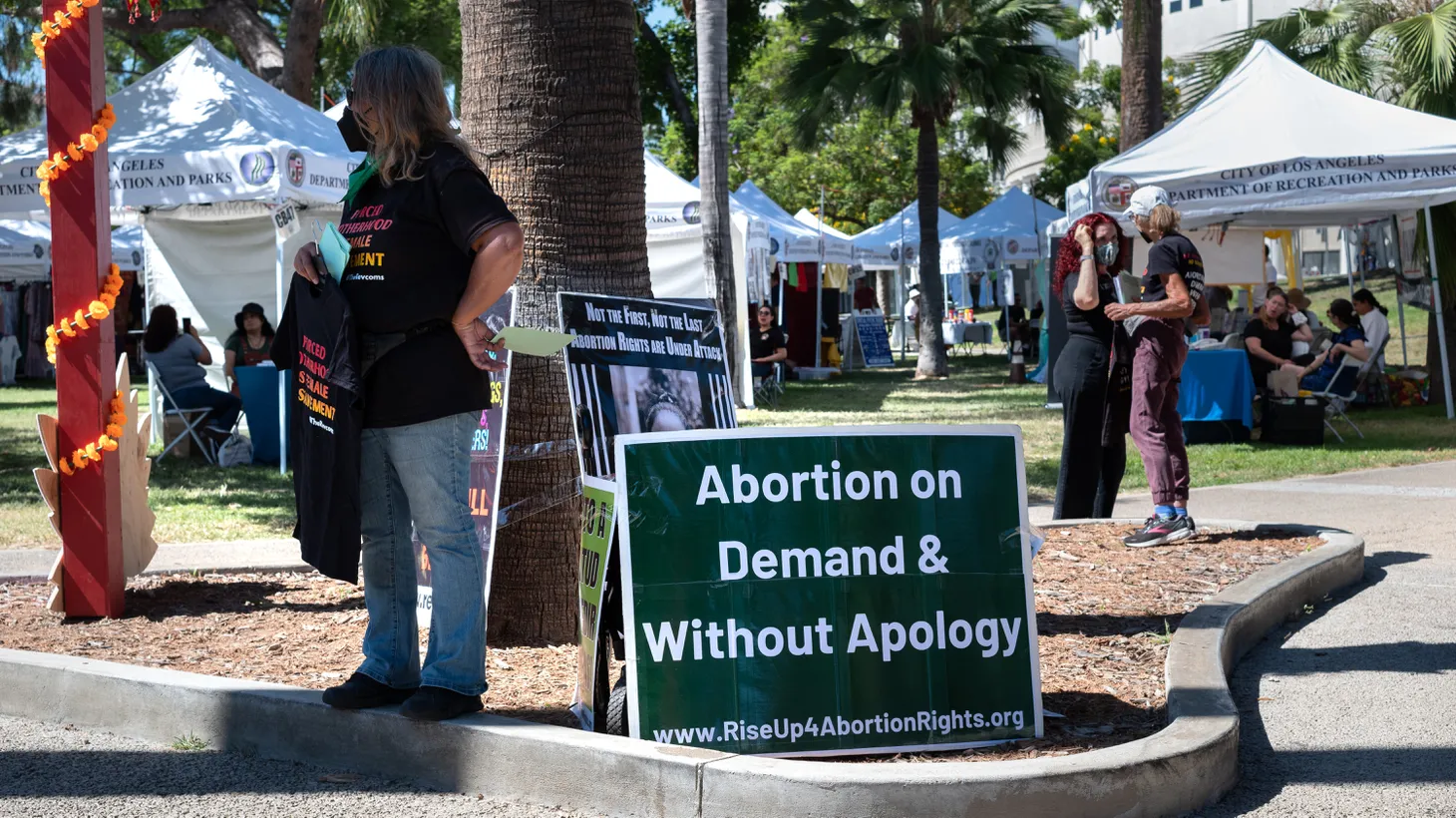 An activist is ready to hand out a t-shirt as she stands next to a sign saying, “Abortion on demand and without apology,” at Echo Park Lake, July 10, 2022.